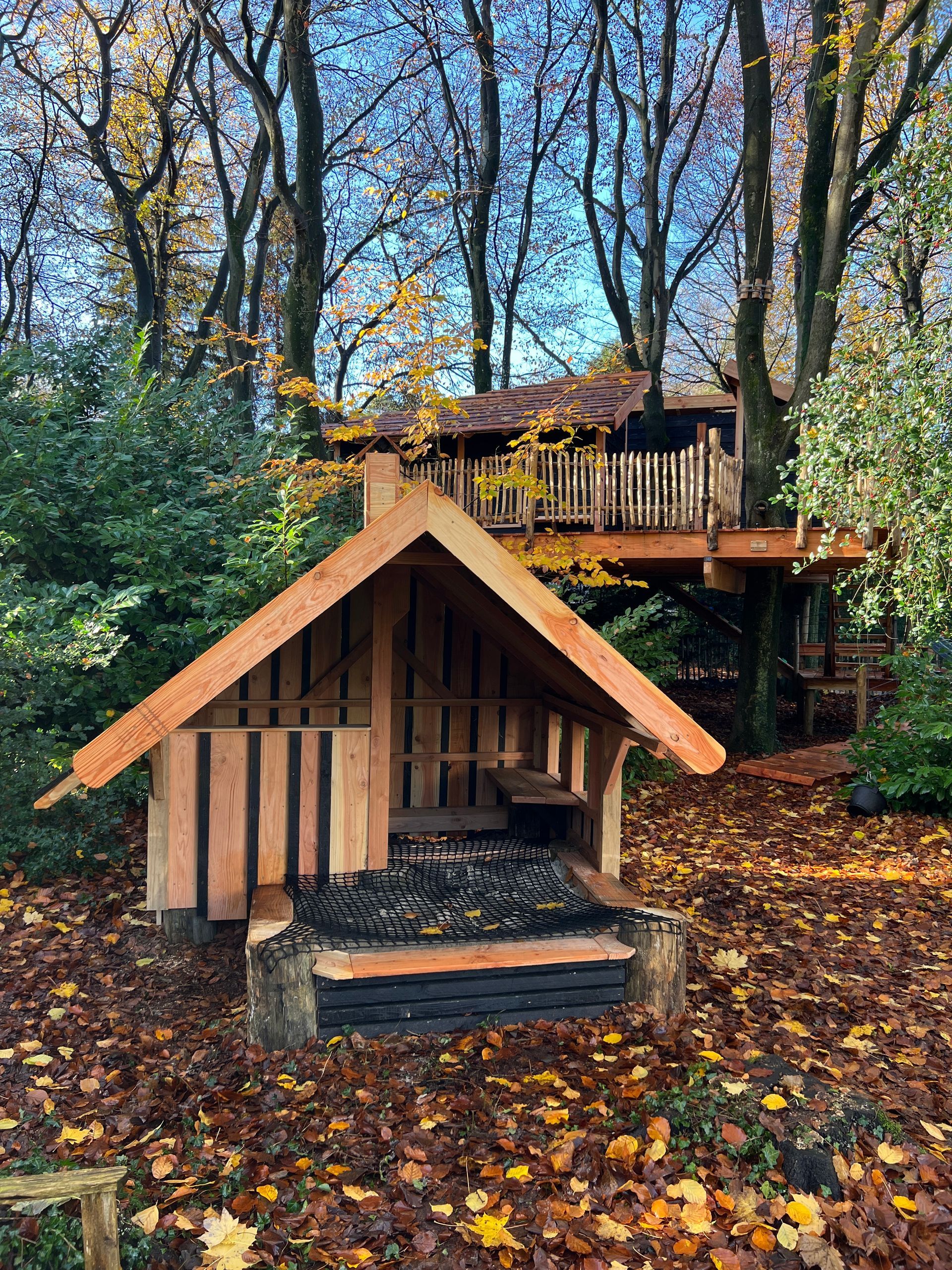Houten speelhuisje met een boomhut op de achtergrond, gelegen in een bos met herfstbladeren.