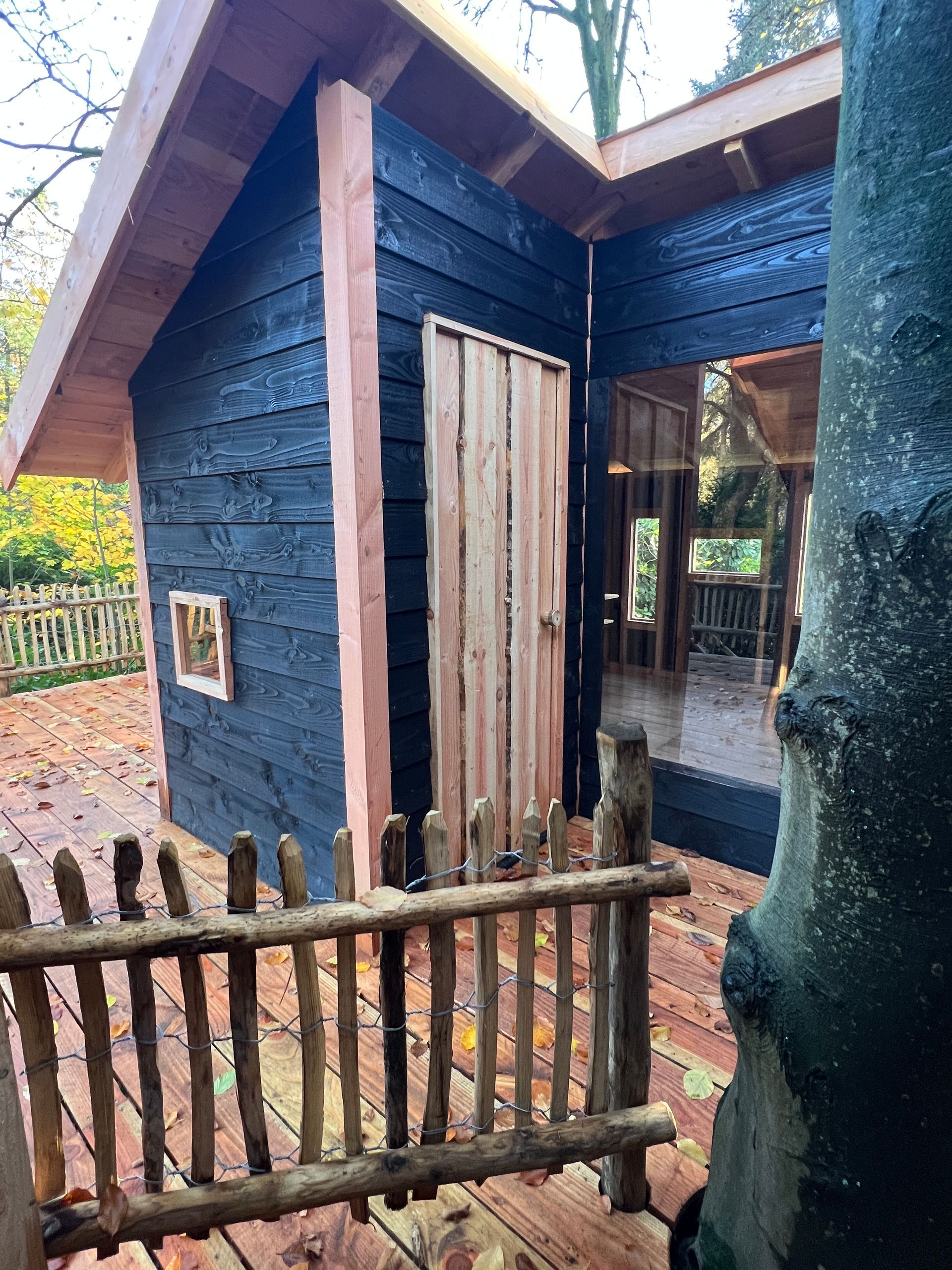 Een zwarte houten boomhut met een rustieke houten schutting en deur.