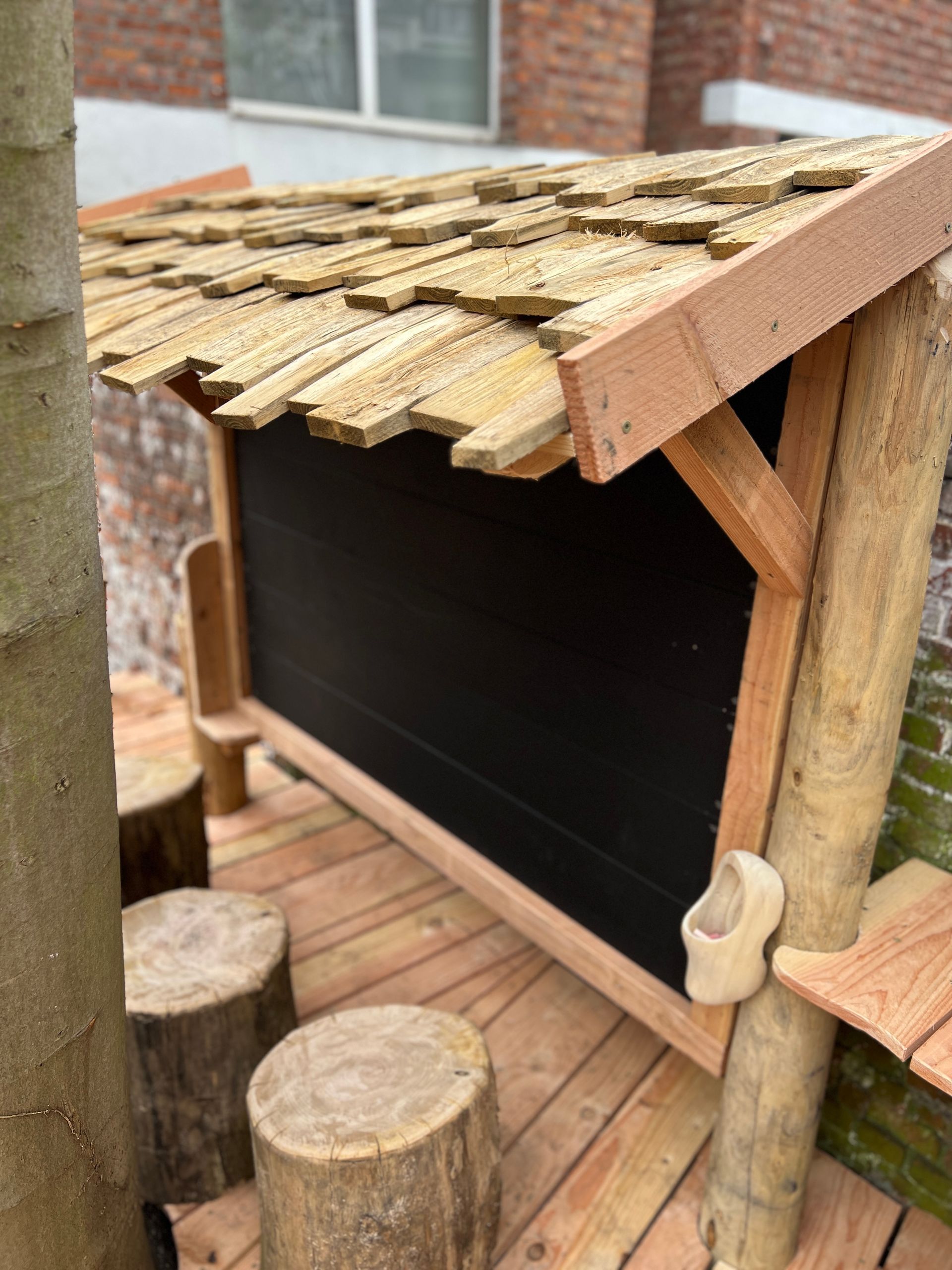 Een houten krijtbord voor buiten met een dak van dakpannen en krukjes van boomstammen, naast een boom.