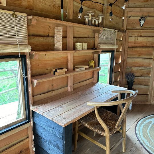 Gezellig interieur van een houten blokhut met een bureau, planken, een stoel en een raam met uitzicht op het groen.