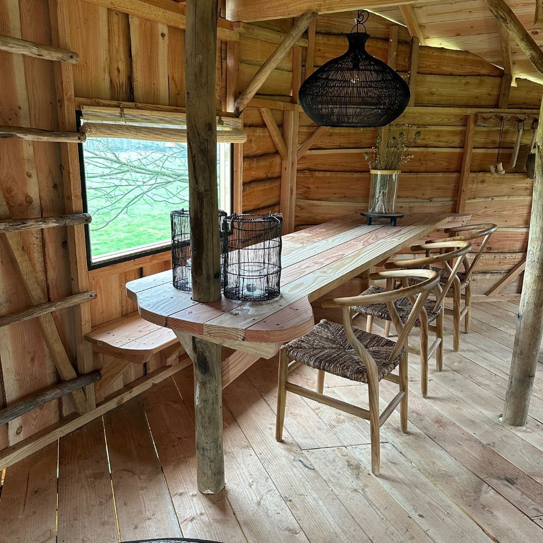 Rustiek houten interieur met een lange tafel, stoelen en een hanglamp. Uitzicht op groen vanuit het raam.