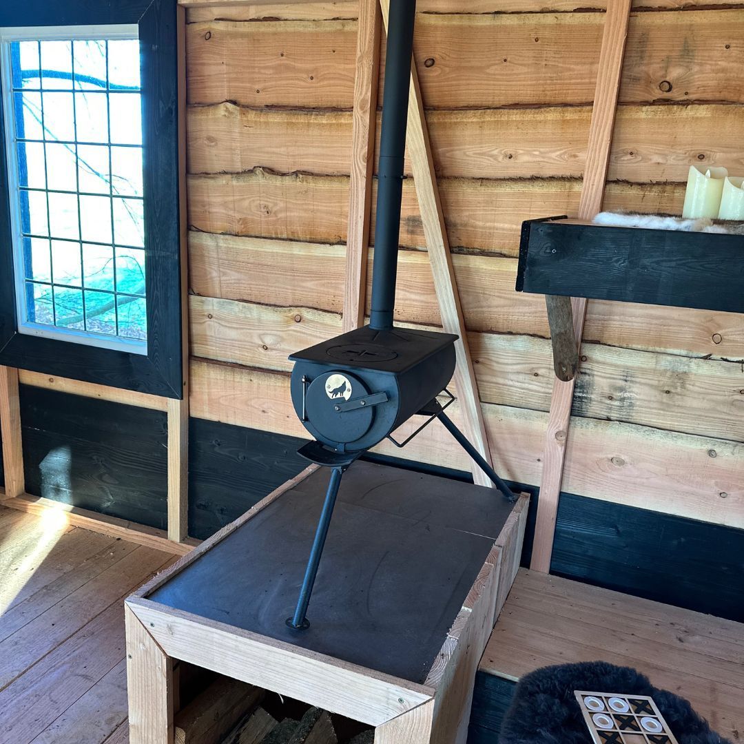 Een kleine houtkachel met schoorsteen in een rustieke houten hut, vlakbij een raam.