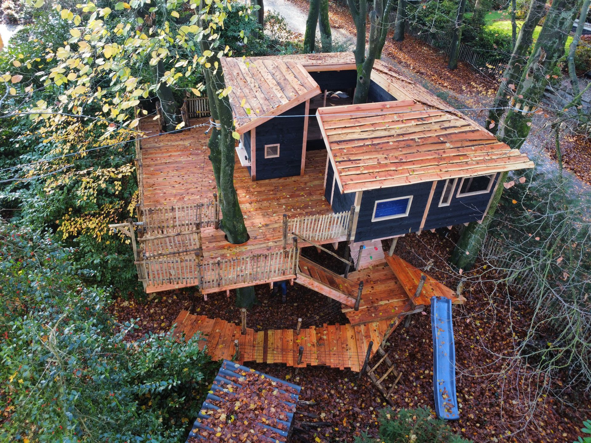 Boomhut met houten platform, meerdere verdiepingen en glijbaan; omgeven door bomen.