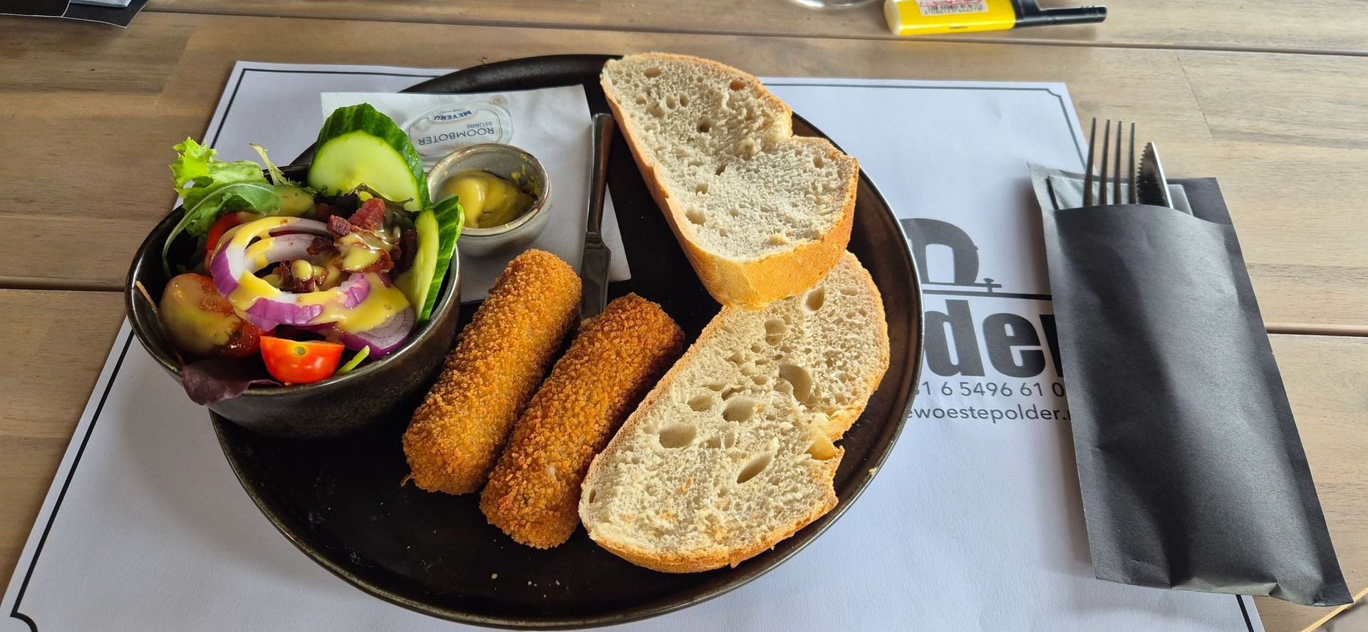 Een bord met salade, brood, gefrituurd eten en mosterd. Een vork en mes liggen naast het bord op een placemat.