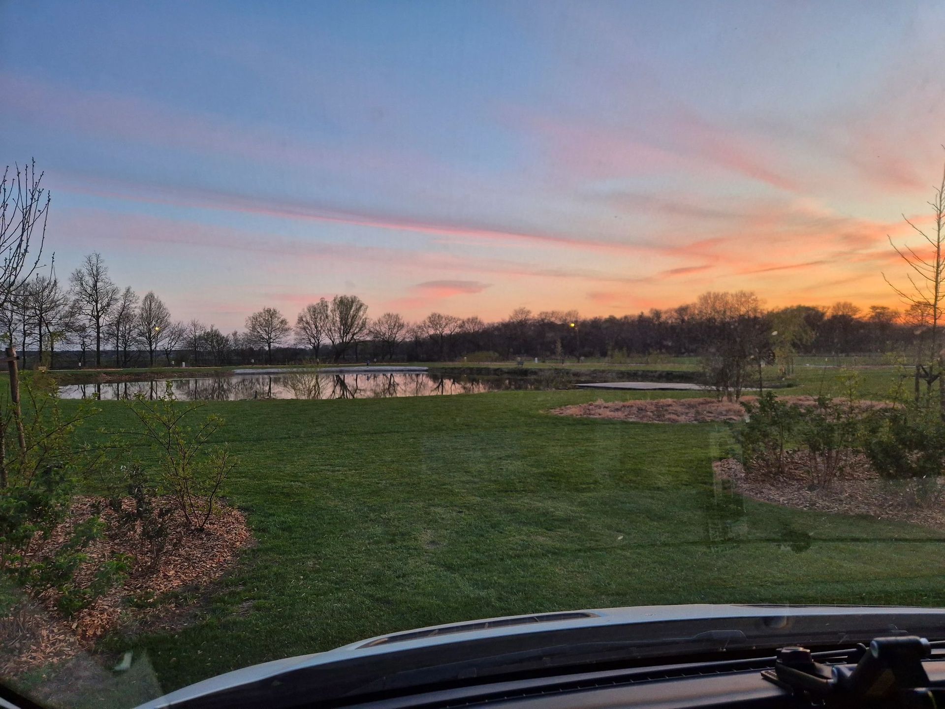 Zonsondergang boven een vijver, groen gras, bomen. Oranje en roze lucht met blauw.