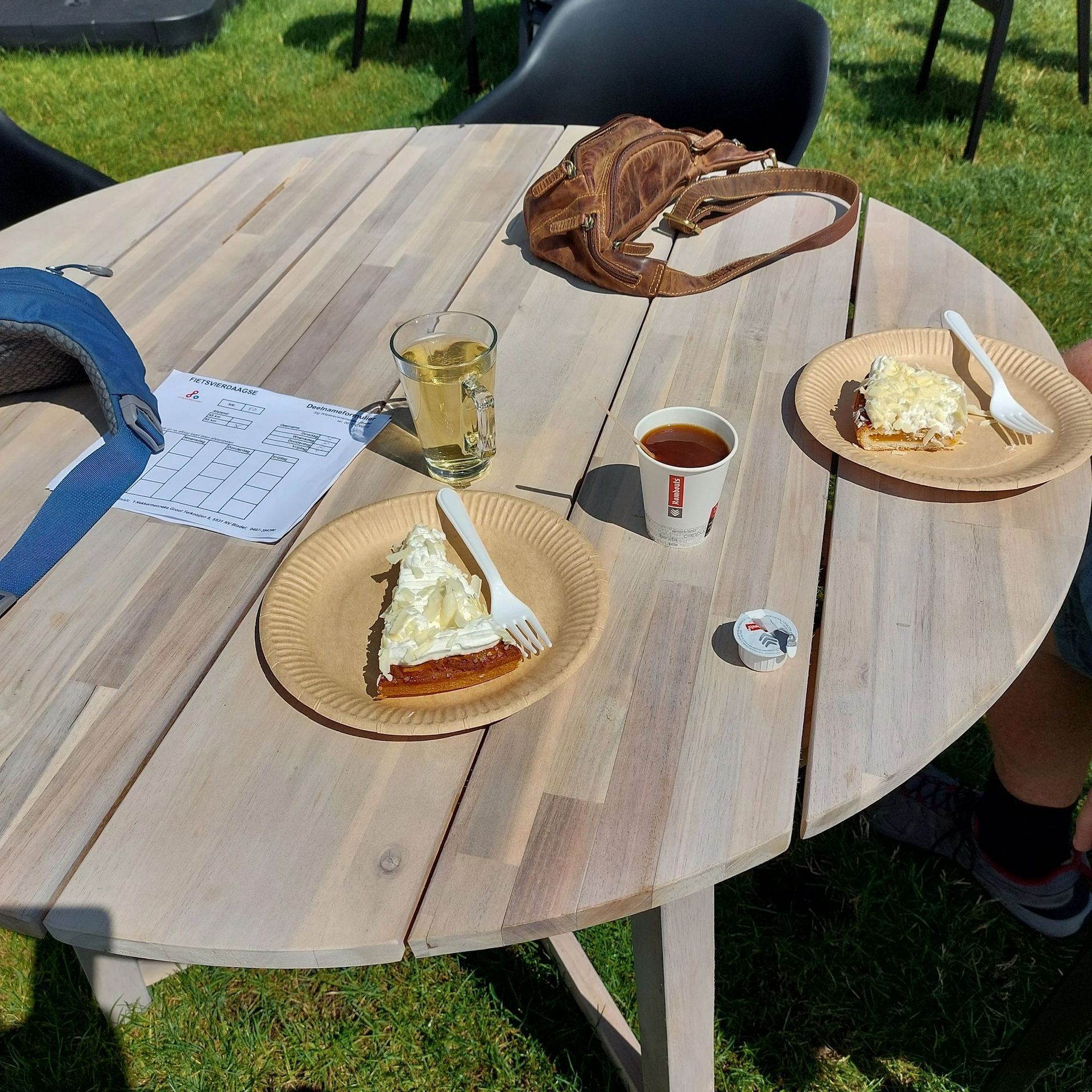 Een houten tafel met eten, drinken, een tas en papieren op een zonnige dag.