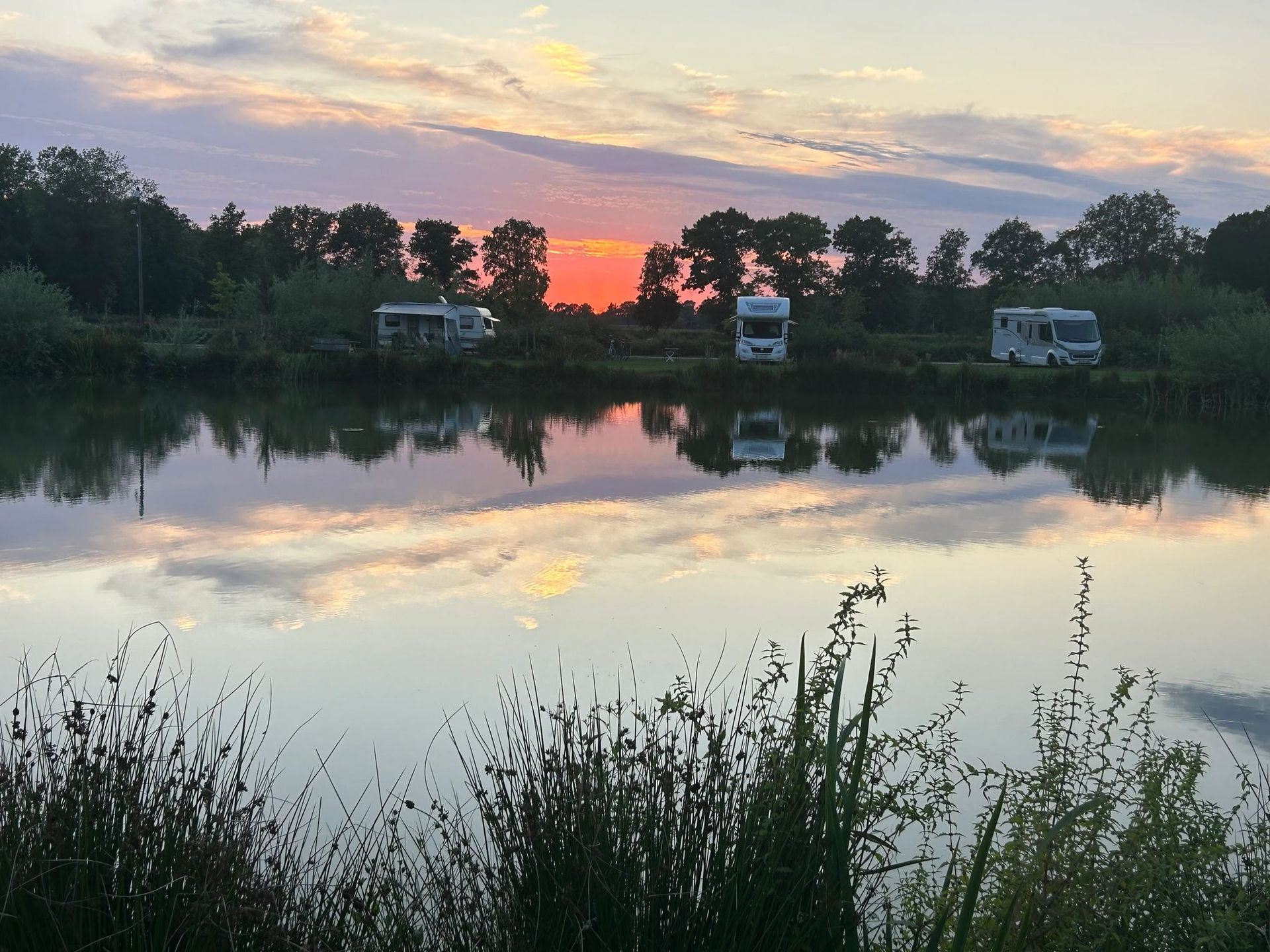 Kampeerders bij een stil meer waarin de kleurrijke zonsondergang wordt weerspiegeld.