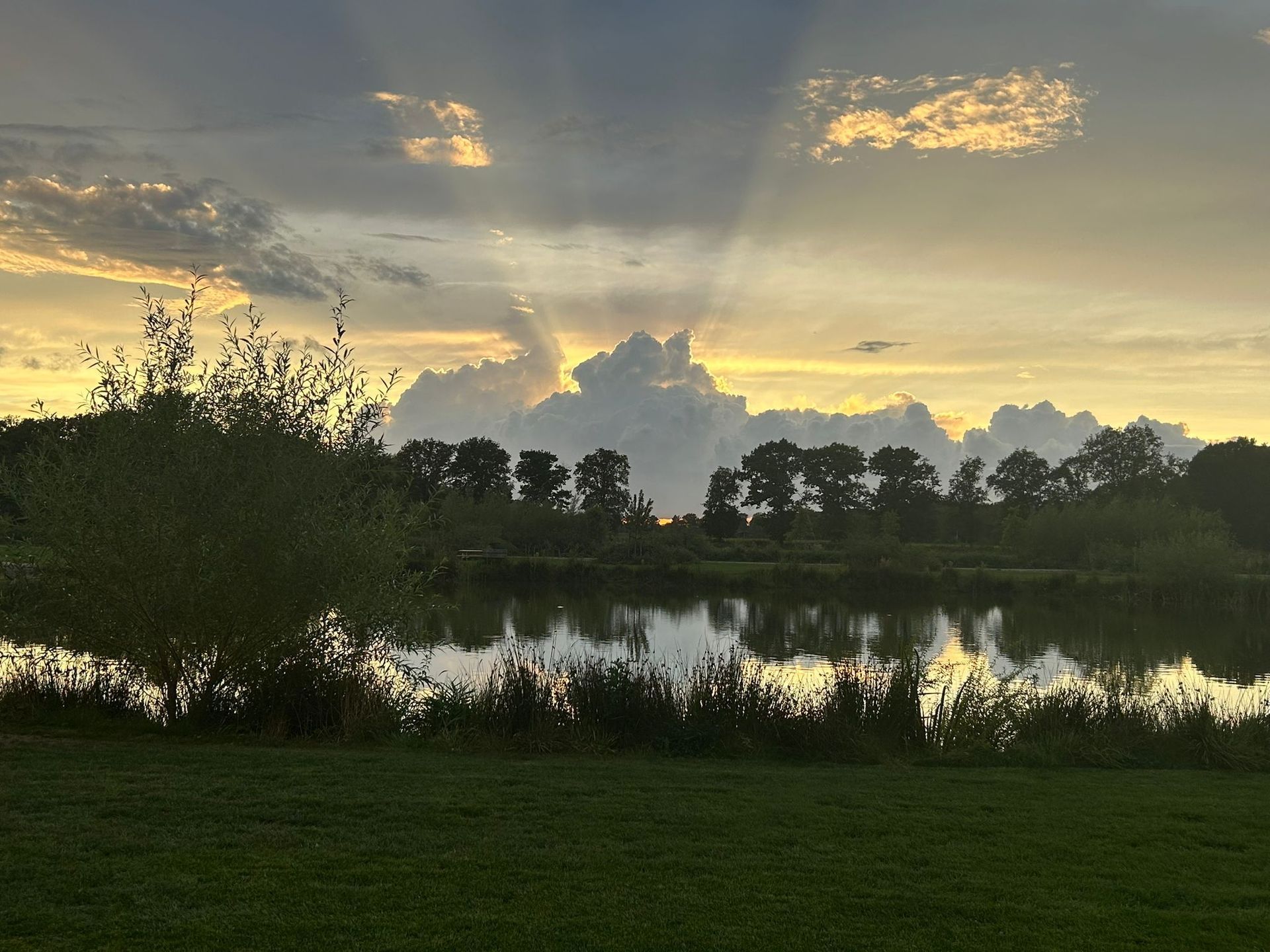 Zonsondergang boven een meer; verlichte wolken die in het water weerspiegelen; bomen aan de overkant van de oever.
