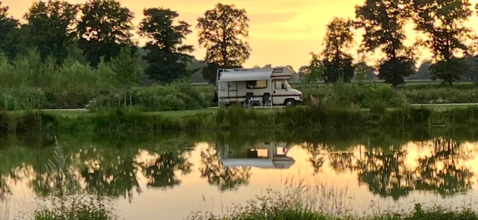 Camper geparkeerd bij een spiegelend water met bomen op de achtergrond tijdens zonsondergang.