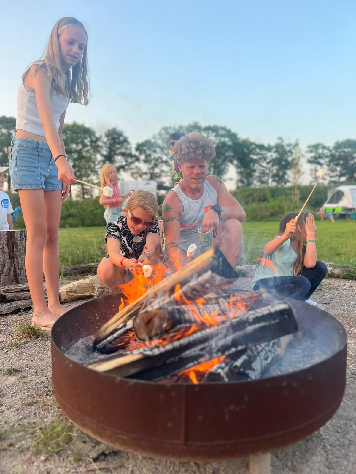 Kinderen roosteren marshmallows boven een kampvuur in een metalen vuurkorf. Groen veld, blauwe lucht.