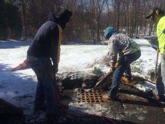 People shoveling snow and debris around a small outdoor fire pit in a snowy wooded area