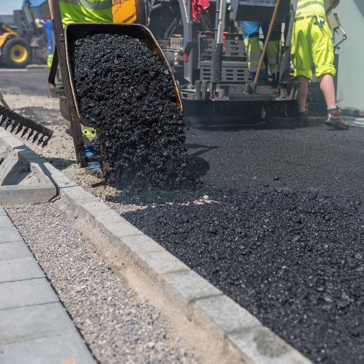 Road paving machine pouring black asphalt beside a curb, with workers in the background.