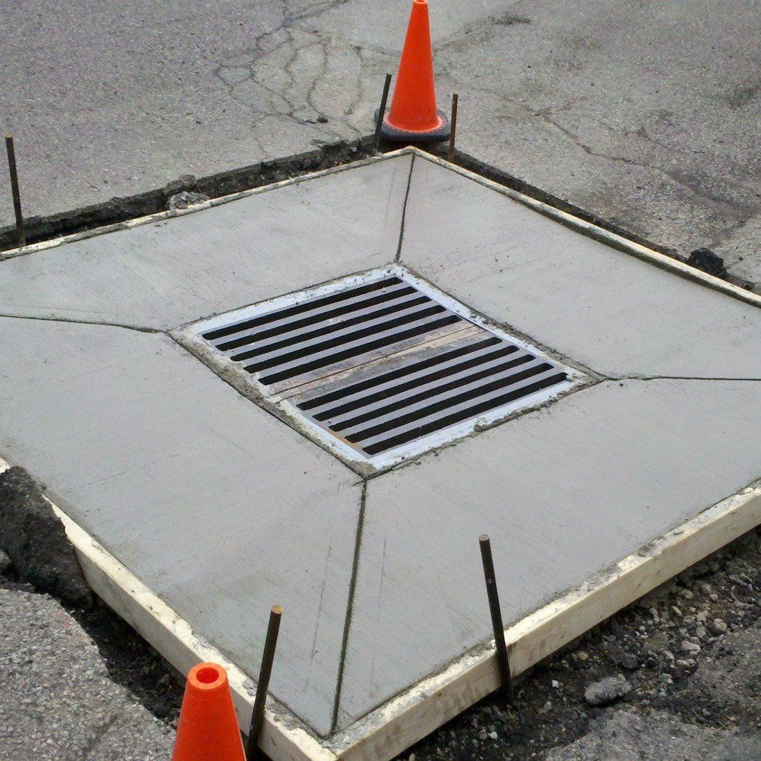 Roadside storm drain with metal grate and orange traffic cones on cracked pavement.