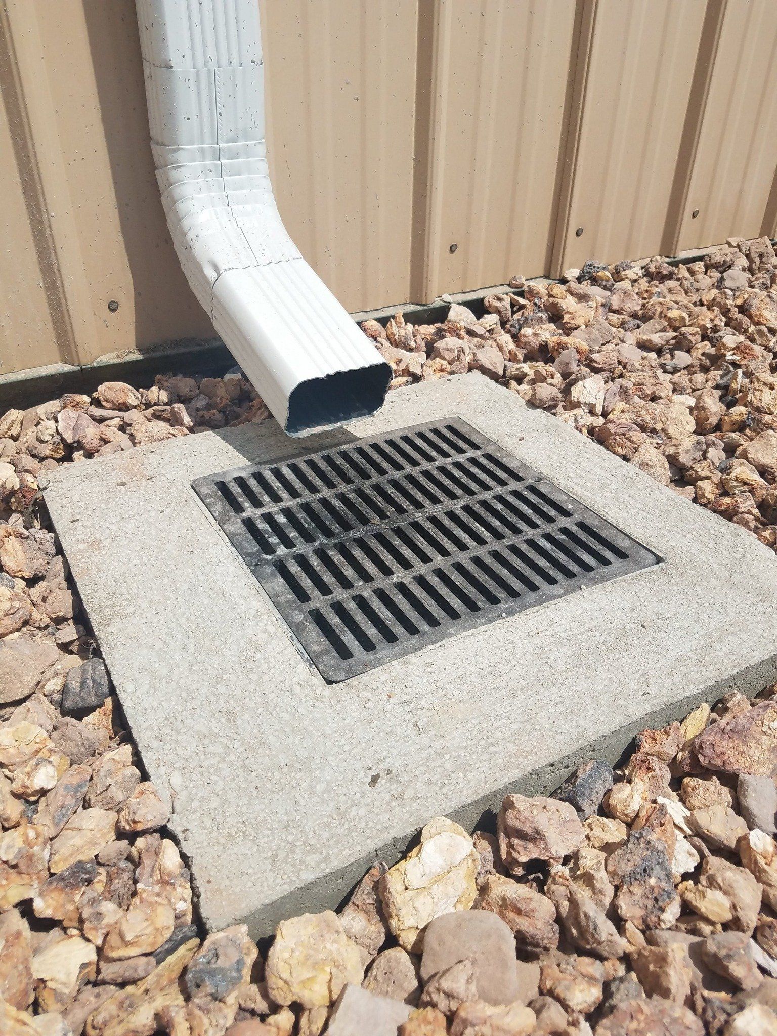 Gutter downspout draining into a concrete storm drain grate beside a tan wall and rocky ground