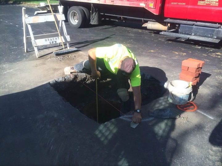 Worker kneeling at an open manhole on a street beside a truck and safety equipment
