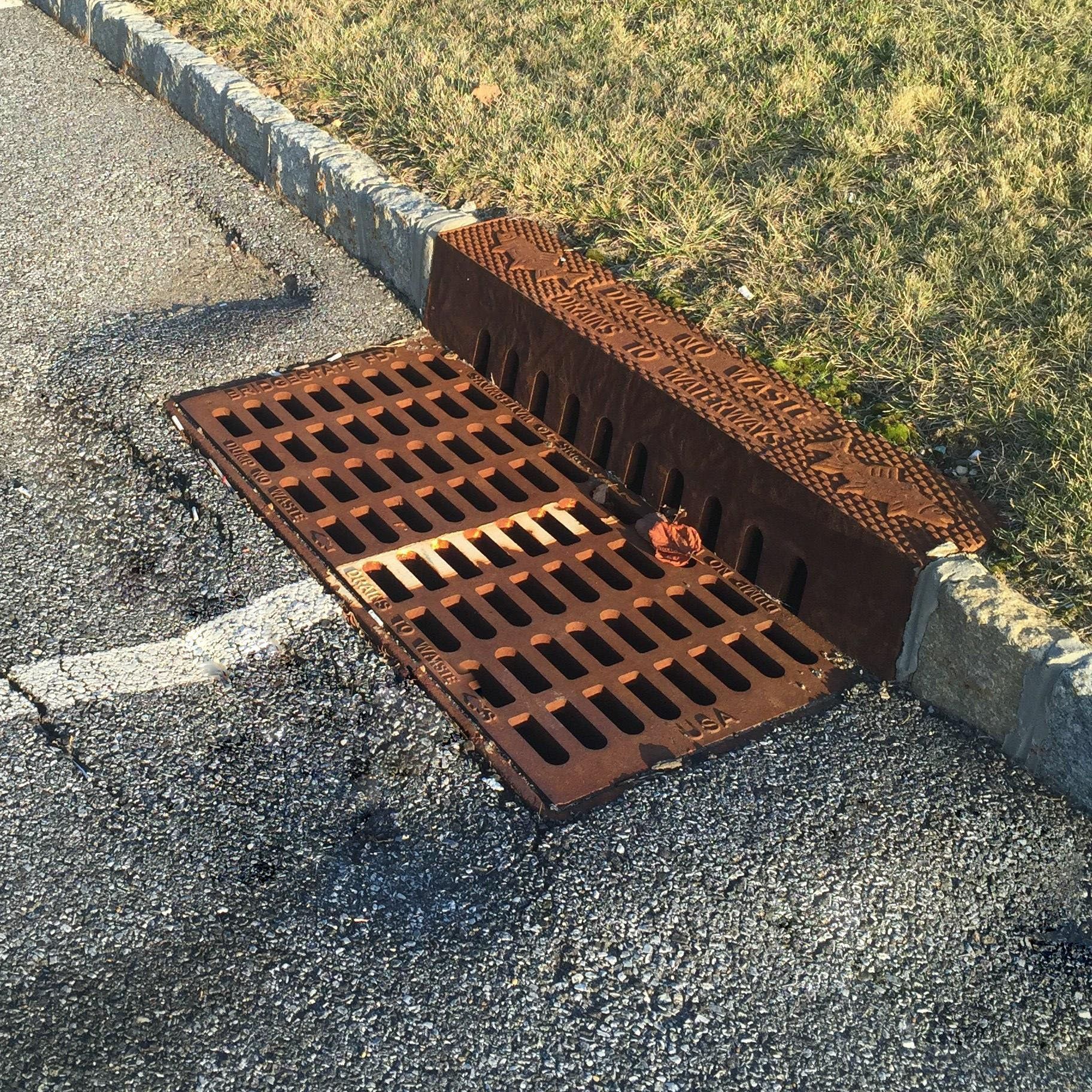 Rusty storm drain grate beside a curb on an asphalt street, with water runoff debris at the edge.