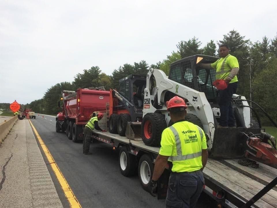 Roadwork crew loading a white skid steer onto a trailer beside a highway, with workers in high-visibility gear