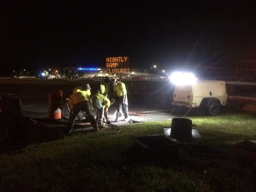 Night roadwork crew in reflective vests repairing pavement beside a bright work truck and cones at night