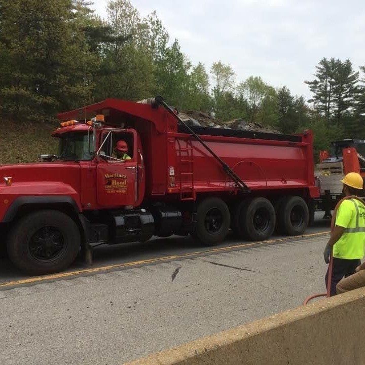 Red dump truck parked on a road work site with a worker in a yellow hard hat nearby.