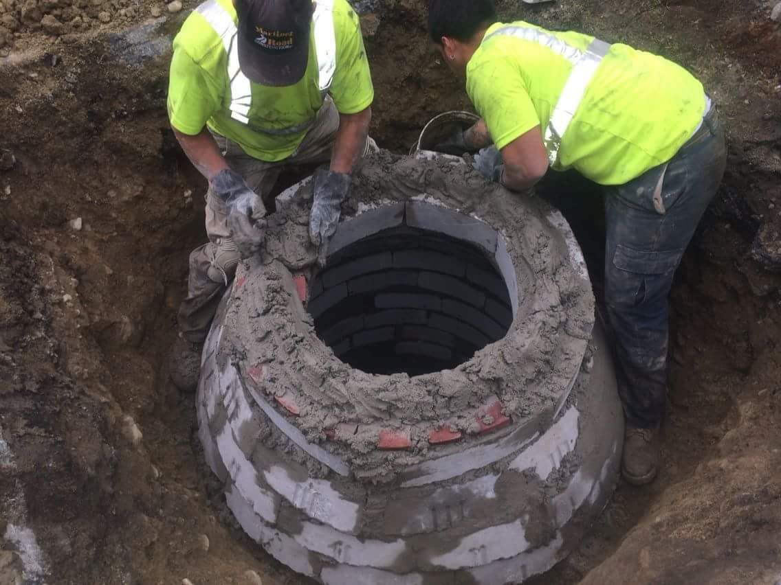 Two workers in neon vests repairing a concrete manhole opening in a dirt trench
