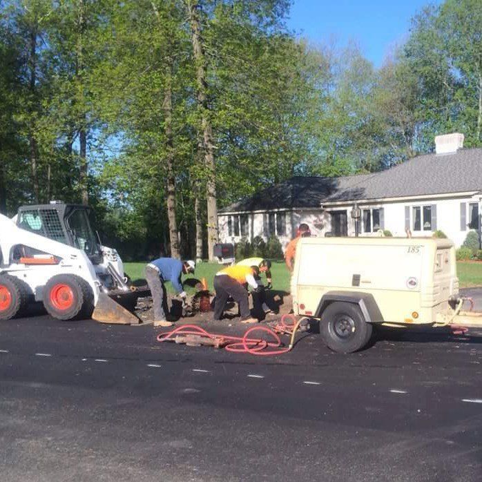 Workers patching a road beside a white skid steer and a cream utility trailer near houses on a sunny day