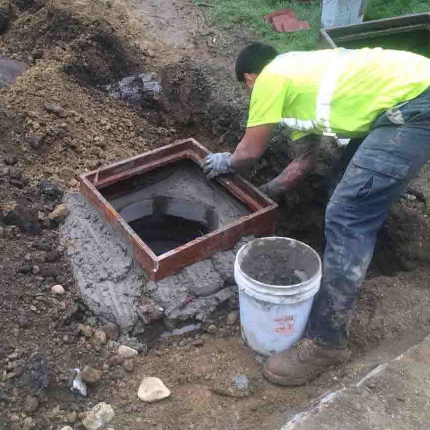 Worker installing a metal utility frame in a dirt trench beside a white bucket and soil pile