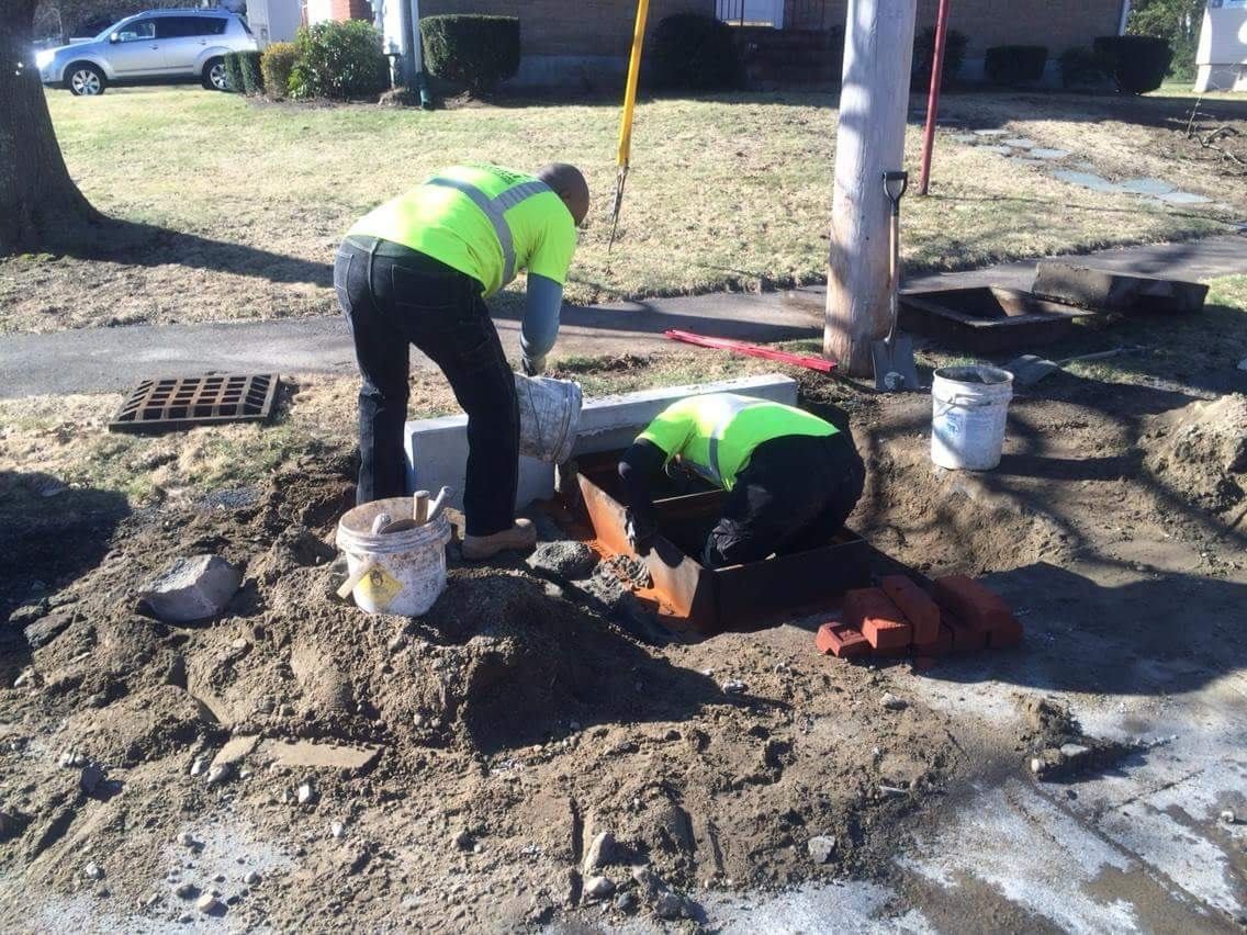 Two workers in neon safety vests repairing a curbside area near a tree with tools and a bucket.