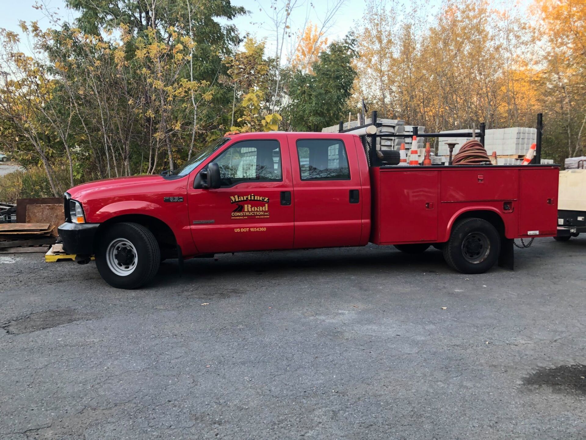 Red utility pickup truck parked on gravel beside autumn trees and equipment trailers