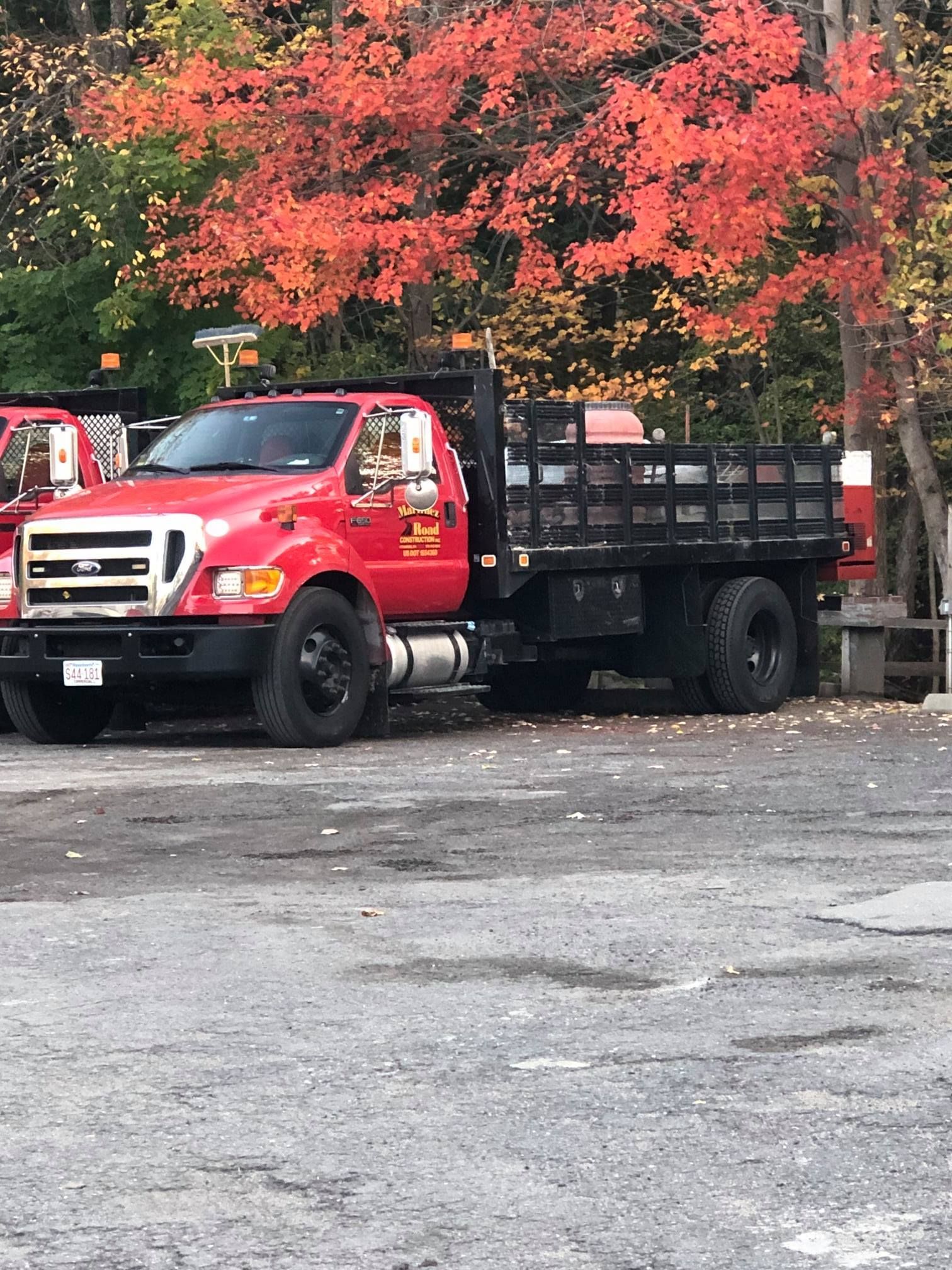 Red flatbed truck parked on a gravel lot beneath bright autumn trees