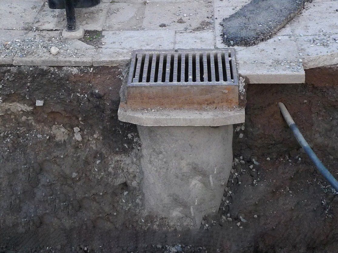 Street drain opening in a concrete curb, with a metal grate and blue hose nearby