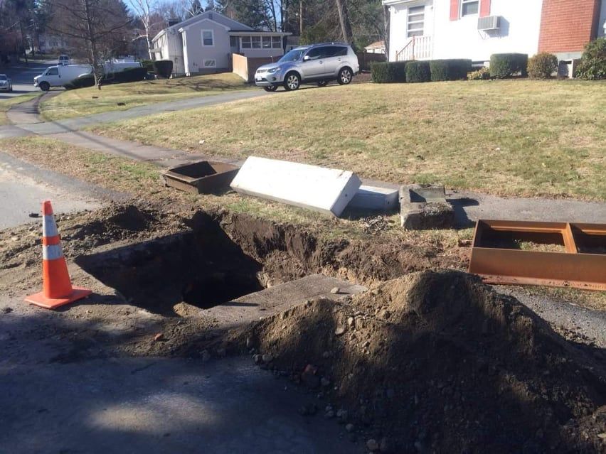 Roadwork excavation trench beside a suburban street, with an orange cone and piled dirt.