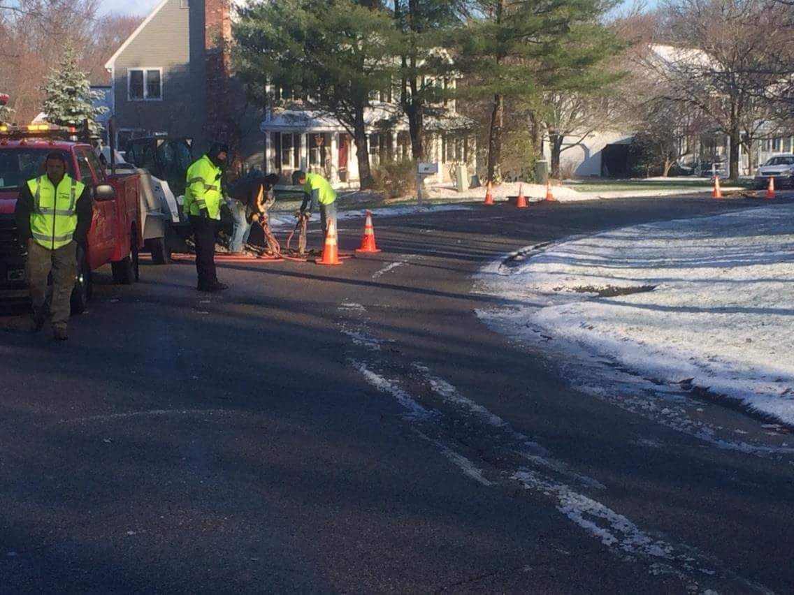 Workers in neon vests set orange cones on a snowy road near a house