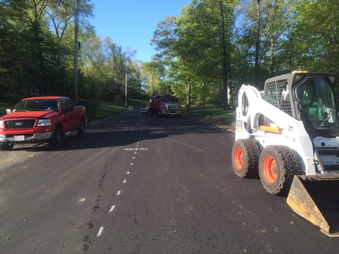 Roadway under construction with a white skid steer and red pickup trucks along a tree-lined street