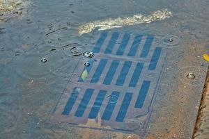 Blue metal storm drain grate in wet pavement with puddles and raindrops