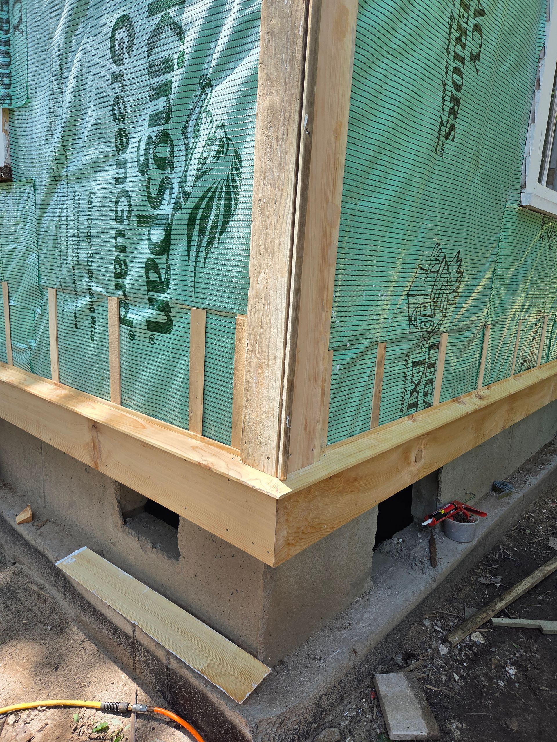 A corner of a building under construction, showing a concrete foundation with ventilation holes, a wooden frame, and wrap.