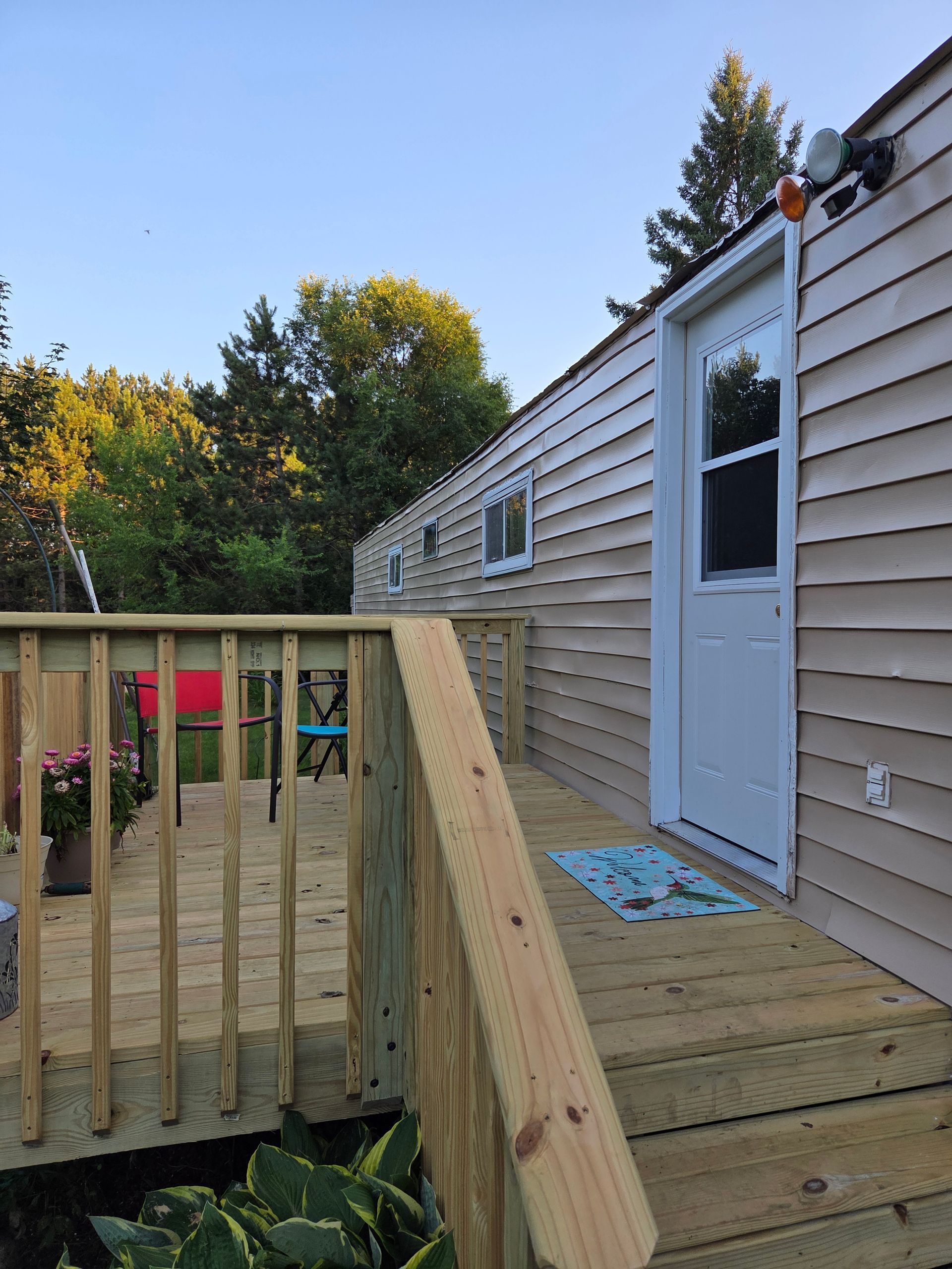 A wooden deck with railings leads to the white door of a light-colored mobile home, set against a backdrop of trees.