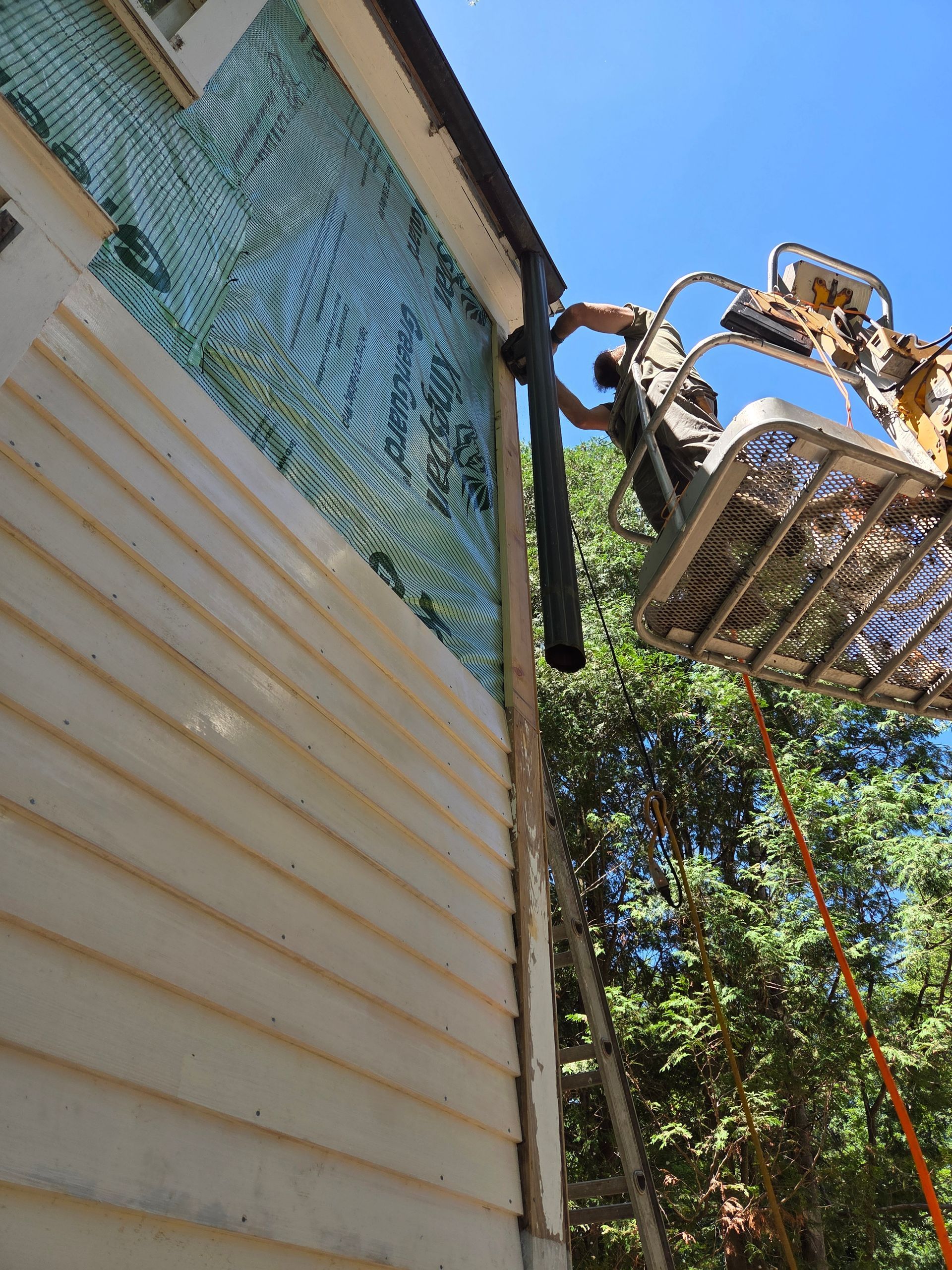 A worker in a boom lift installs a black downpipe on the side of a building under renovation.
