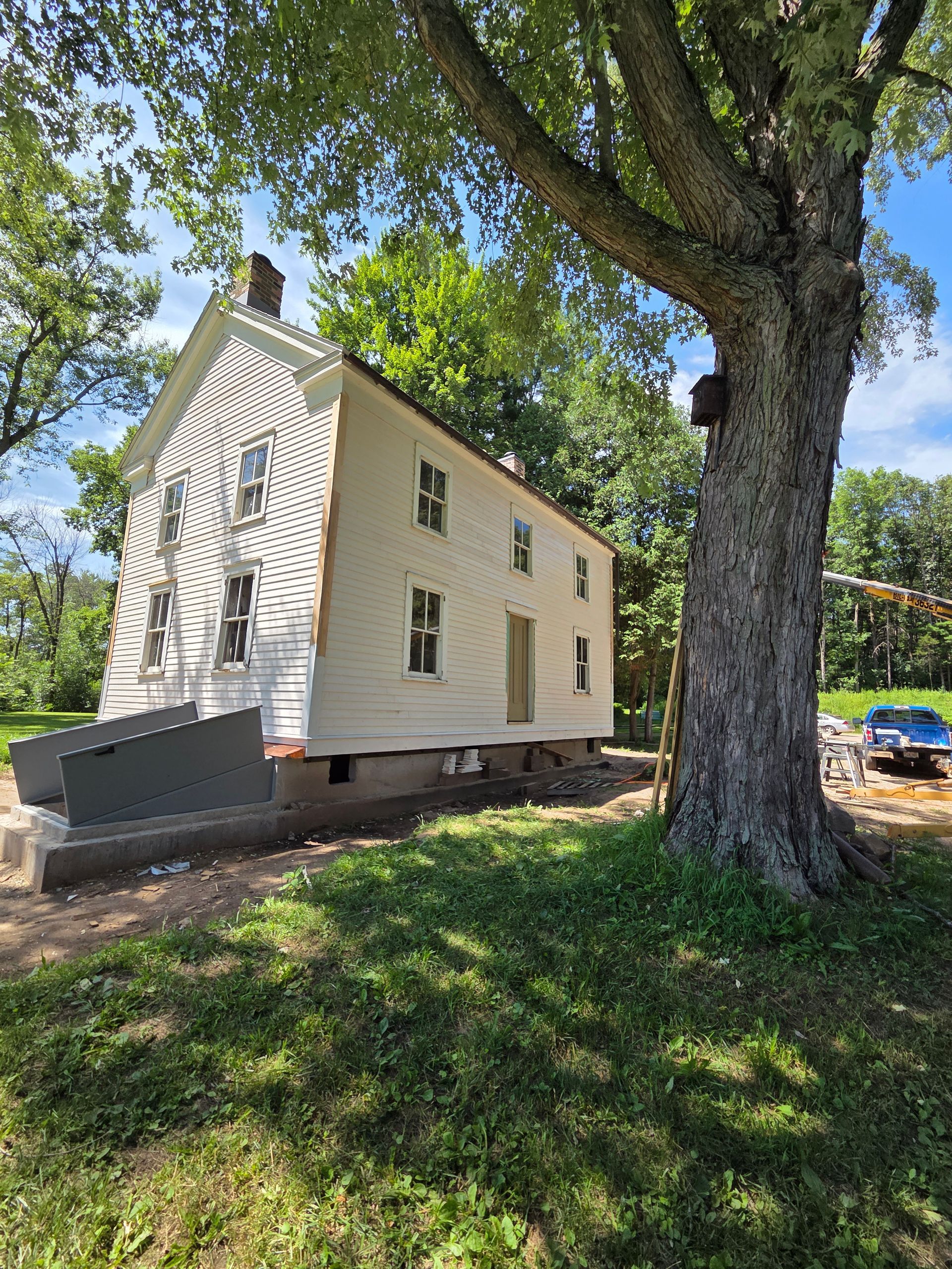 A white, two-story house under renovation, raised on a new foundation, next to a large tree on a sunny day.