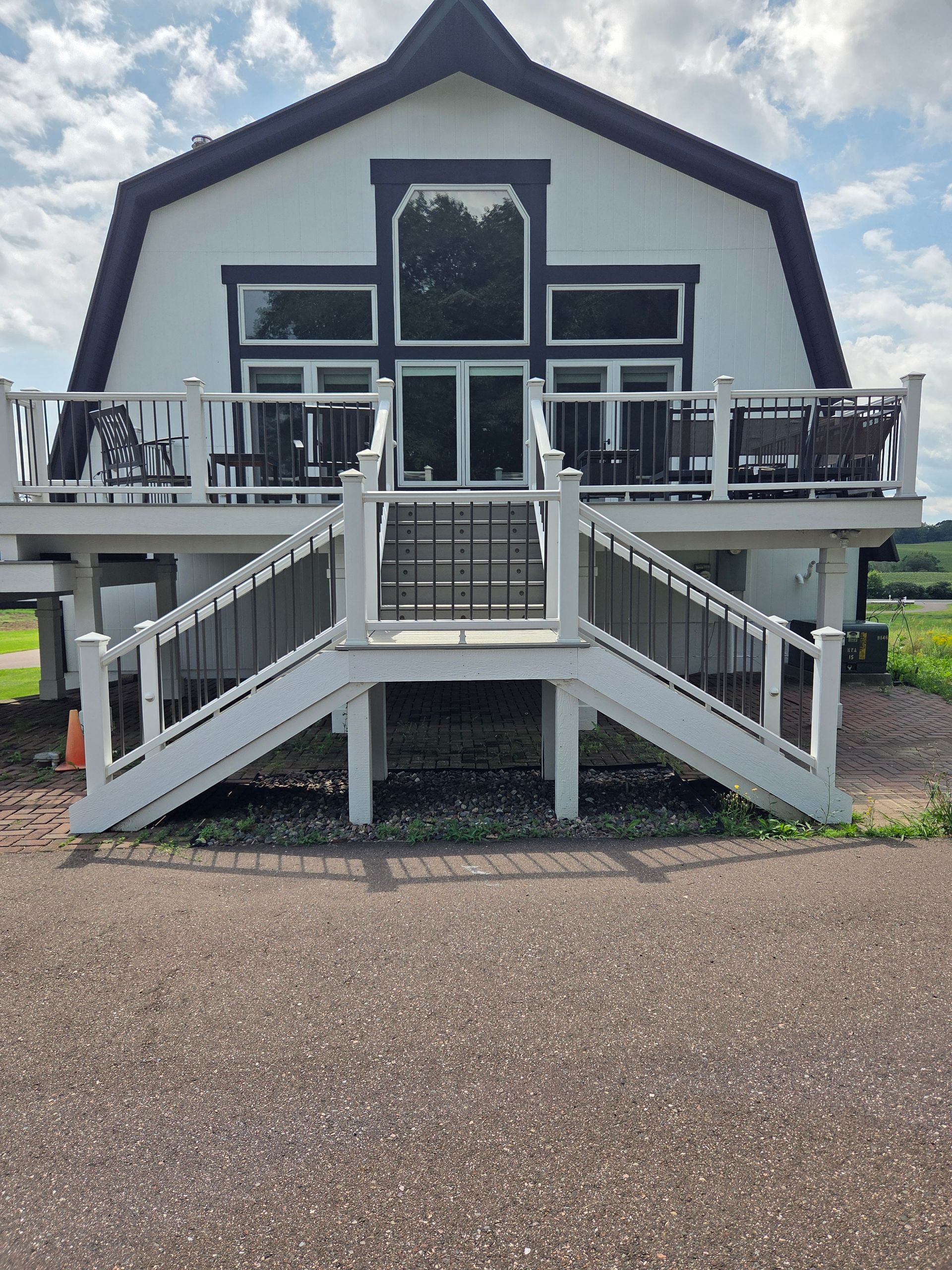 A white, barn-style building with a large deck and dual-staircase entrance over a gravel driveway under a cloudy sky.