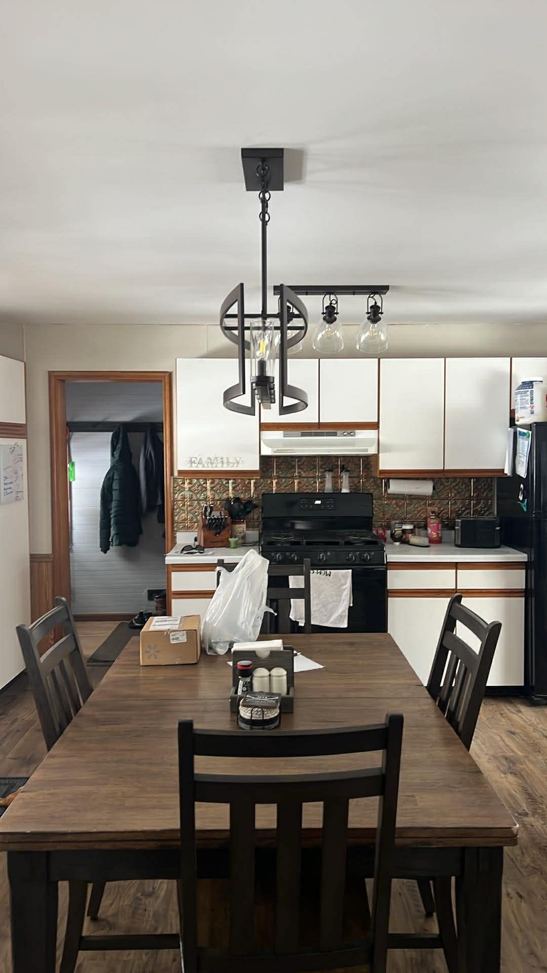 A dining table set in front of a white kitchen with wooden cabinets, a dark pendant light, and black appliances.