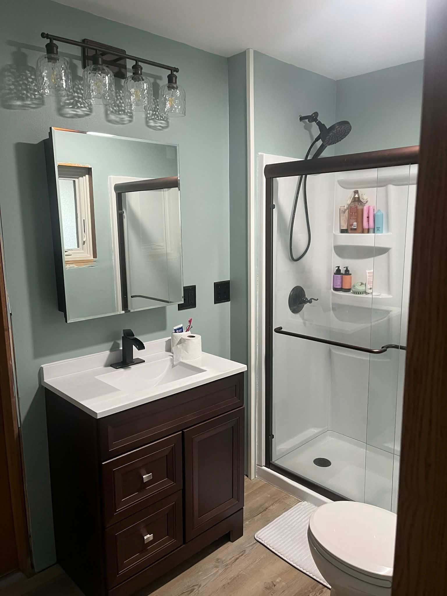 A modern bathroom with sage green walls, a dark wood vanity, a glass-enclosed shower, and a light-toned wood floor.