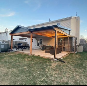 Covered backyard patio with a wooden roof, seating area, and string lights beside a house at dusk Covered backyard patio with a wooden roof, seating area, and string lights beside a house at dusk
