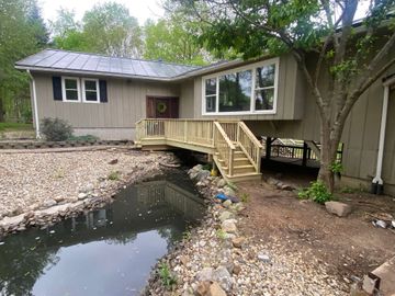 Single-story house with a small wooden porch over a rocky creek or pond in a wooded yard Single-story house with a small wooden porch over a rocky creek or pond in a wooded yard