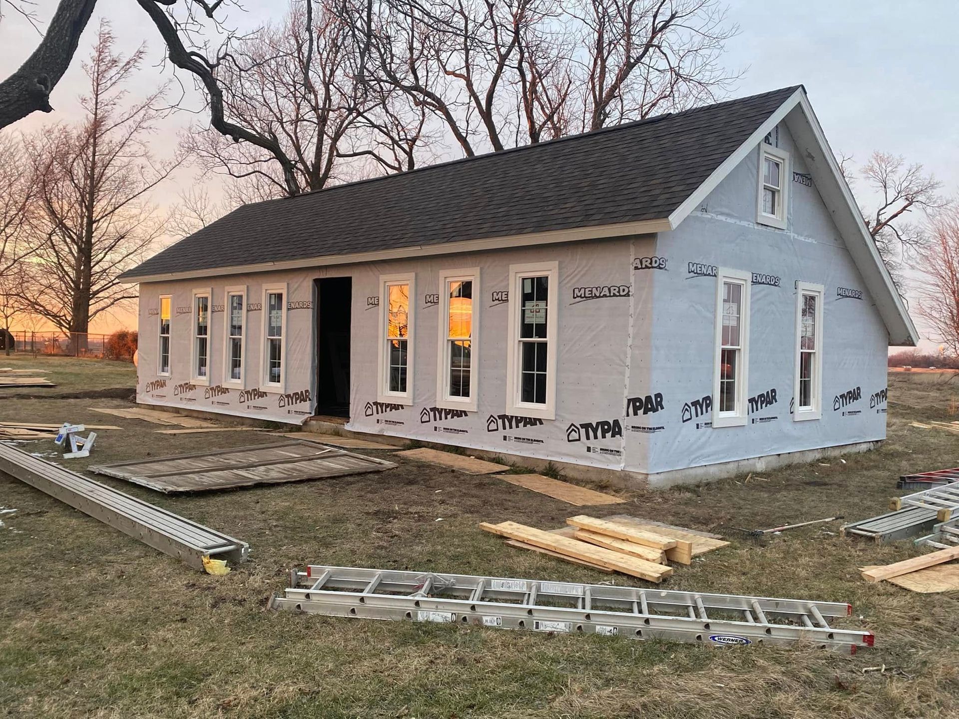 Partially built small house with white siding, black roof, and windows at sunset on a construction site Partially built small house with white siding, black roof, and windows at sunset on a construction site