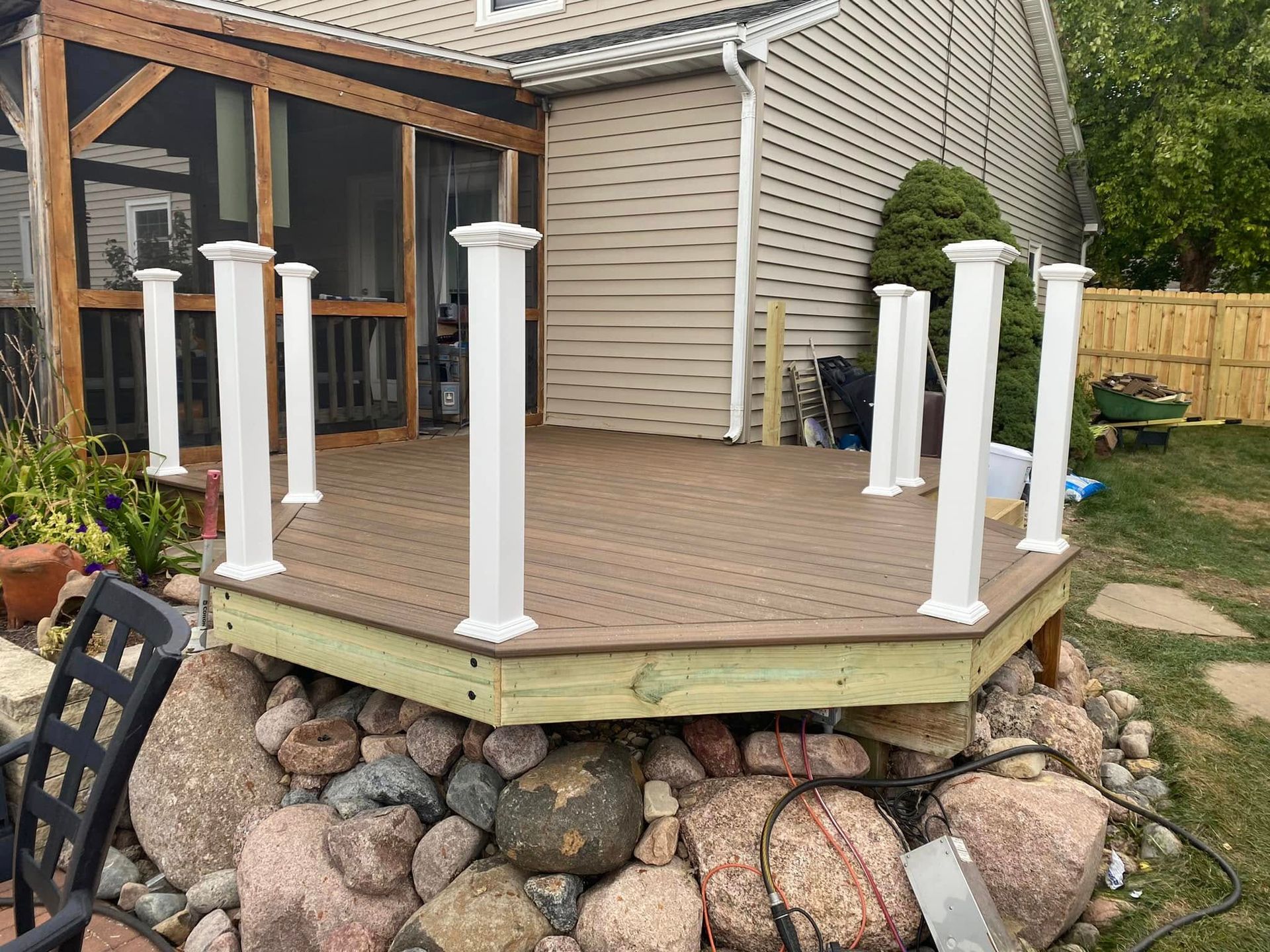 Back deck with white rail posts and stone retaining wall beside a house, surrounded by trees and shrubs