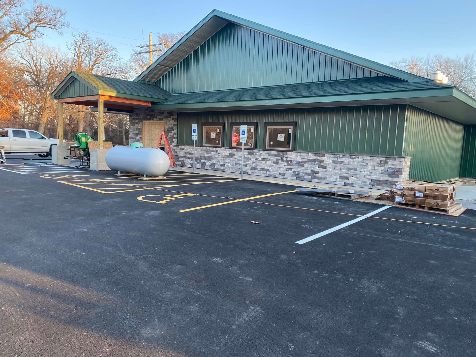 Green-roofed roadside building with stone base, windows, and a propane tank in front, beside an empty parking lot