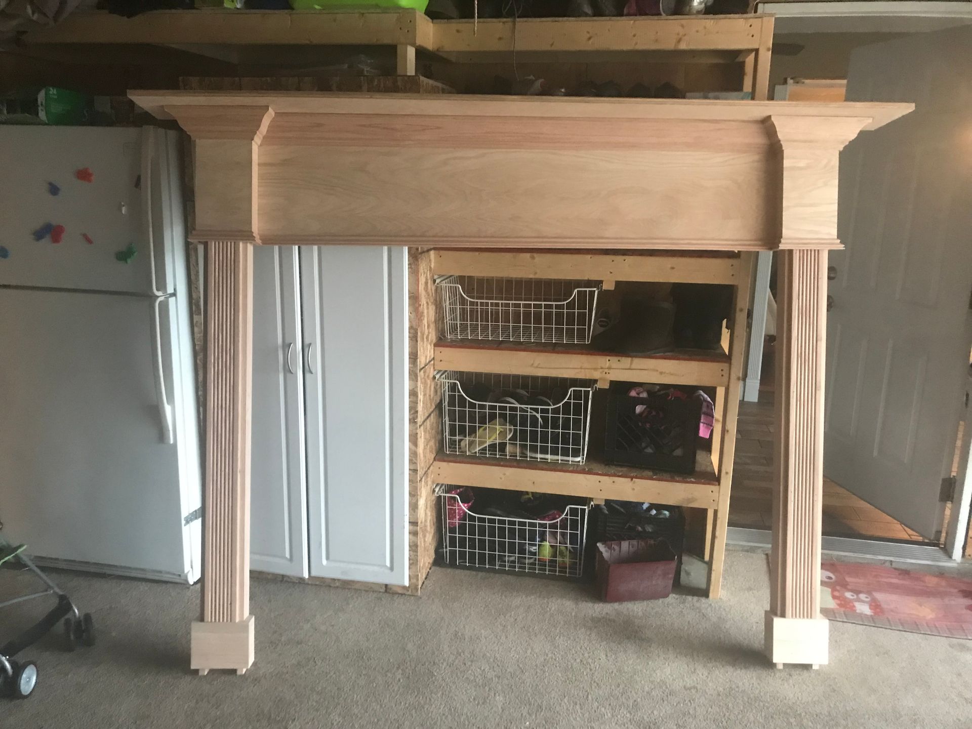 Wooden fireplace mantel with shelves and drawers in a room, with baskets and a refrigerator nearby