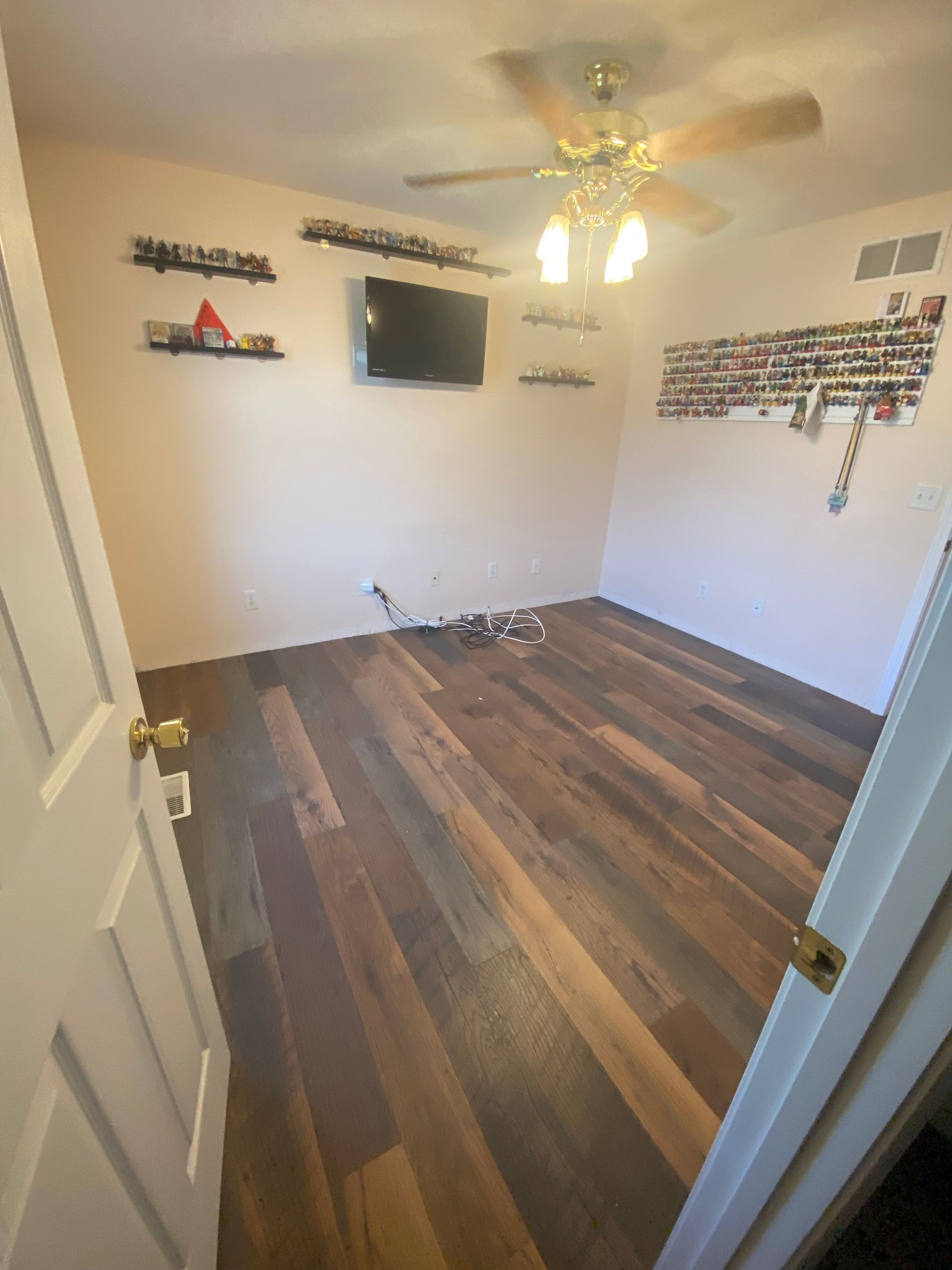 Empty bedroom with wood-look flooring, white walls, ceiling fan, wall-mounted TV, and shelves.