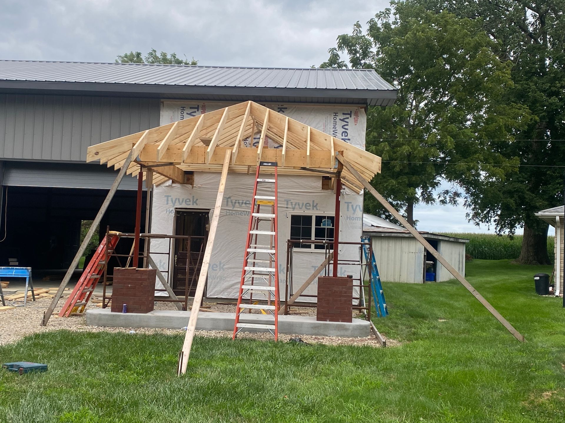 Front porch under construction with exposed framing, ladder, tools, and siding on a sunny day. Front porch under construction with exposed framing, ladder, tools, and siding on a sunny day.