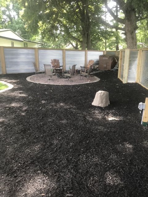 Backyard patio with chairs and table on a circular gravel area, surrounded by a white fence and trees.